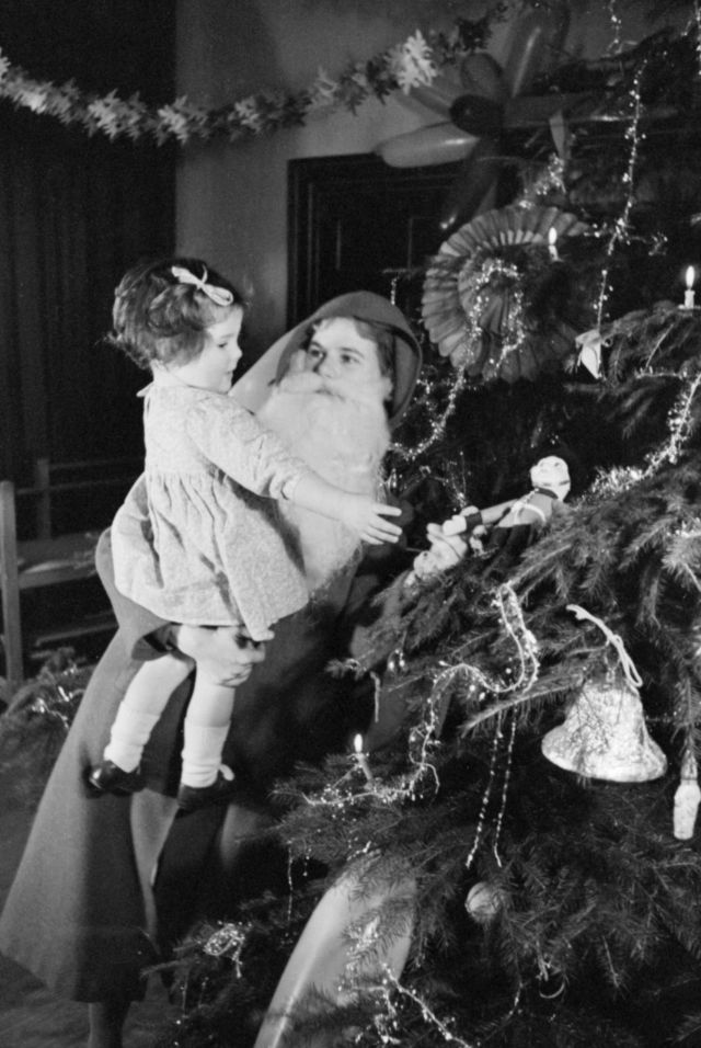 weiblicher Weihnachtsmann mit Kind auf dem Arm und dekorierter Weihnachtsbaum schwarz weiß